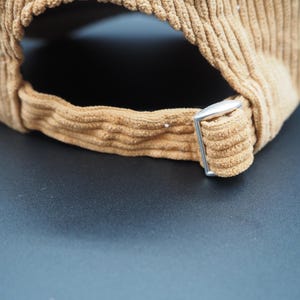 K&ouml;nnte beinhalten: Nahaufnahme einer beigefarbenen Cord-Baseballkappe. Der verstellbare Riemen hat eine silberne Metallschnalle. Der Stoff der Kappe hat eine gerippte Textur. Die Kappe ist neutralfarben.