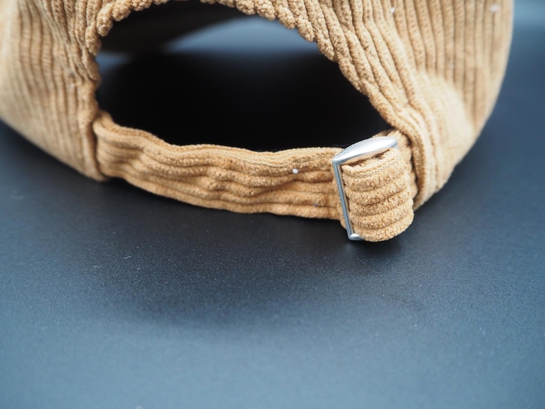 K&ouml;nnte beinhalten: Nahaufnahme einer beigefarbenen Cord-Baseballkappe. Der verstellbare Riemen hat eine silberne Metallschnalle. Der Stoff der Kappe hat eine gerippte Textur. Die Kappe ist neutralfarben.