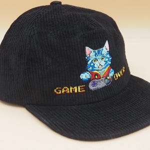 May include: Black corduroy baseball cap featuring an embroidered design of a blue cat holding a video game controller. The words "GAME OVER" are embroidered in yellow. A stylish accessory for gamers.