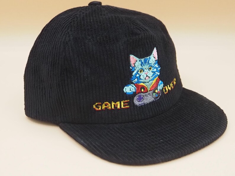 May include: Black corduroy baseball cap featuring an embroidered design of a blue cat holding a video game controller. The words "GAME OVER" are embroidered in yellow. A stylish accessory for gamers.
