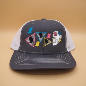 May include: A gray trucker hat with a white mesh back and a curved brim. The front features embroidered designs of birds, triangles, and a skull in various colors. The hat has a snapback closure.