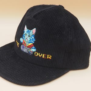 May include: Black corduroy baseball cap with an embroidered design of a blue cat playing a video game. The cat is wearing a red and yellow shirt. The words "GAME OVER" are embroidered in yellow.