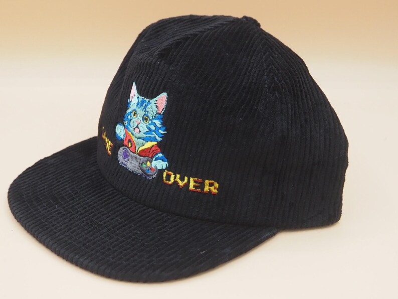 May include: Black corduroy baseball cap with an embroidered design of a blue cat playing a video game. The cat is wearing a red and yellow shirt. The words "GAME OVER" are embroidered in yellow.