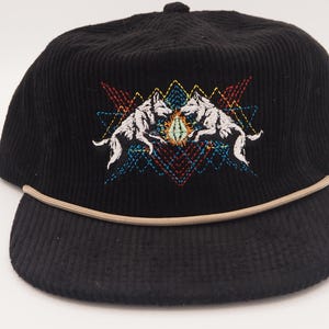 May include: Black corduroy cap with a curved brim and a tan rope detail. The cap features an embroidered design of two white wolves facing each other, set against a geometric pattern with red, blue, and yellow accents.