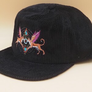 K&ouml;nnte beinhalten: Schwarze Cord-Baseballkappe mit einem gestickten Design, das zwei gefl&uuml;gelte Fabelwesen in Orange, Rosa und T&uuml;rkis zeigt. Die Kappe hat einen gebogenen Schirm und eine strukturierte Krone.