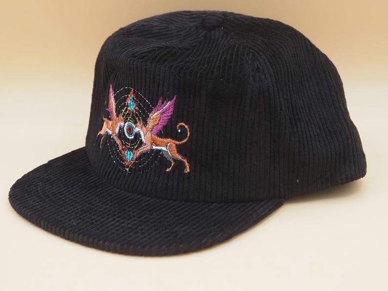 K&ouml;nnte beinhalten: Schwarze Cord-Baseballkappe mit einem gestickten Design, das zwei gefl&uuml;gelte Fabelwesen in Orange, Rosa und T&uuml;rkis zeigt. Die Kappe hat einen gebogenen Schirm und eine strukturierte Krone.
