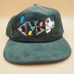 May include: Dark green corduroy baseball cap with a flat brim. The cap features embroidered designs including a skull, birds, and geometric shapes in various colors. A black rope detail accents the brim.