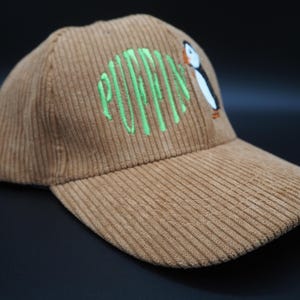 K&ouml;nnte beinhalten: Braune Cord-Baseballkappe mit dem gr&uuml;n gestickten Wort "PUFFINS" und einem Cartoon-Papageientaucher. Die Kappe hat einen gebogenen Schirm und eine strukturierte Krone.