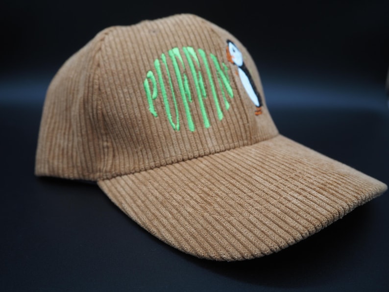 K&ouml;nnte beinhalten: Braune Cord-Baseballkappe mit dem gr&uuml;n gestickten Wort "PUFFINS" und einem Cartoon-Papageientaucher. Die Kappe hat einen gebogenen Schirm und eine strukturierte Krone.