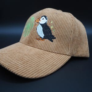 K&ouml;nnte beinhalten: Beige Cord-Baseballkappe mit gesticktem Papageientaucher-Design. Der Papageientaucher ist schwarz, wei&szlig; und orange, mit gr&uuml;nen Akzenten. Die Kappe hat einen gebogenen Schirm und eine strukturierte Krone.
