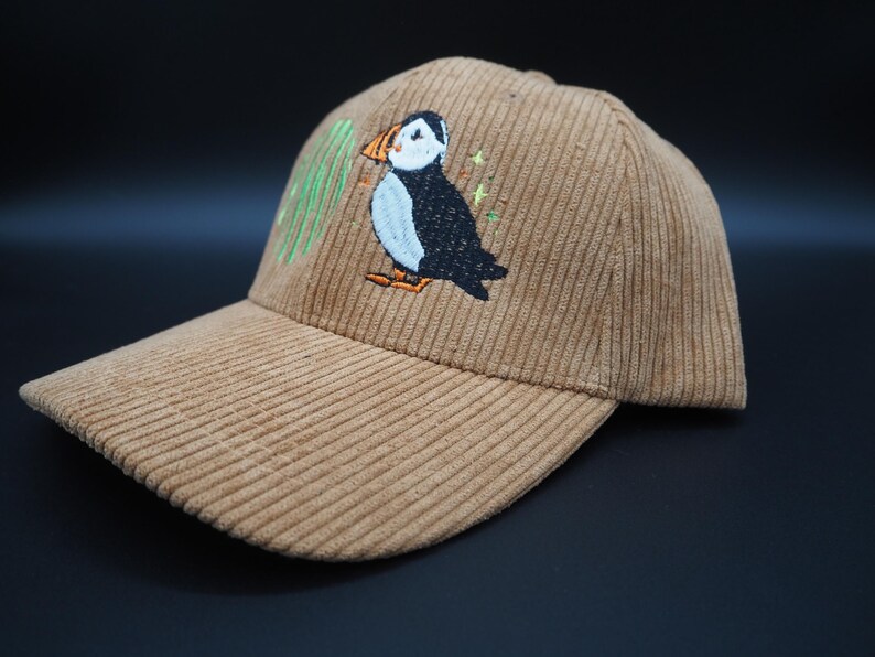 K&ouml;nnte beinhalten: Beige Cord-Baseballkappe mit gesticktem Papageientaucher-Design. Der Papageientaucher ist schwarz, wei&szlig; und orange, mit gr&uuml;nen Akzenten. Die Kappe hat einen gebogenen Schirm und eine strukturierte Krone.
