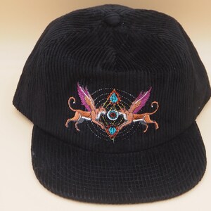 K&ouml;nnte beinhalten: Schwarze Cord-Baseballkappe mit einem bestickten Design. Das Design zeigt zwei gefl&uuml;gelte Kreaturen mit rosa Fl&uuml;geln und einem geometrischen Muster. Die Kappe hat einen gebogenen Schirm.
