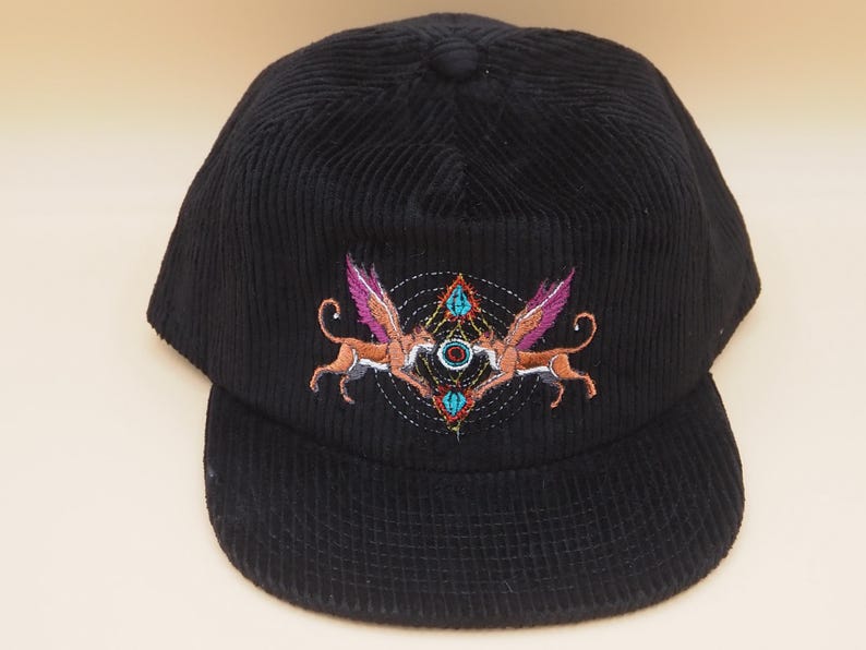 K&ouml;nnte beinhalten: Schwarze Cord-Baseballkappe mit einem bestickten Design. Das Design zeigt zwei gefl&uuml;gelte Kreaturen mit rosa Fl&uuml;geln und einem geometrischen Muster. Die Kappe hat einen gebogenen Schirm.