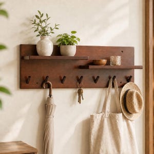 Op de afbeelding: Een donkerbruine houten wandorganizer met twee horizontale planken en zeven zwarte metalen haken. Op de planken staan twee groene planten in pot, een kleine houten kom en een glazen waxinelichthouder. Een paraplu, sleutels, een tas en een hoed hangen eronder.