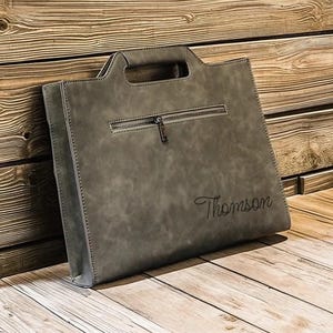 Peut inclure: Sac fourre-tout en cuir gris avec une poignée supérieure et une poche zippée. Le sac porte le nom "Thomson" écrit en cursive. Le sac est fait d'un matériau texturé et est posé sur une surface en bois.