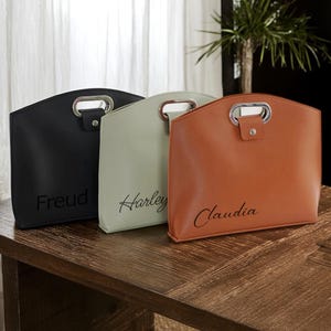 Könnte beinhalten: Drei personalisierte Tragetaschen aus schwarzem, salbeigrünem und hellbraunem Leder. Jede Tasche hat einen silbernen Griff, einen Druckknopfverschluss und einen Namen: Freud, Harley und Claudia. Die Taschen stehen auf einer Holzoberfläche.
