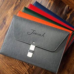 May include: A selection of document holders in shades of grey, red, green, orange, and blue. The grey holder is personalised with the name "Josiah". Each holder has a flap closure with a metal clasp and a white strap.