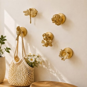 May include: A collection of five gold-toned wall hooks featuring detailed animal and floral motifs, including a dragonfly, a dog head, a bird, a goldfish, and a flower. One hook holds a beige woven tote bag filled with white daisies.