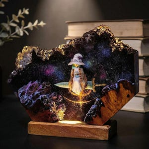 May include: A decorative lamp featuring a miniature figure of a witch riding a sword, set against a galaxy backdrop. The lamp is encased in a dark, textured resin with gold accents, mounted on a wooden base. The lamp emits a warm, inviting glow.