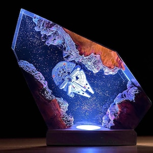 May include: A unique resin art piece featuring a Millennium Falcon model set against a starry background. The artwork is encased in a clear, angled resin block, with textured rock formations in shades of red, purple, and brown. The base is a dark wood.