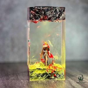 May include: A resin sculpture featuring a miniature warrior figure with a red cape and gold accents, standing on a green and red landscape. The sculpture is encased in a clear, rectangular block with a dark, textured top. The base has a yellow glow.