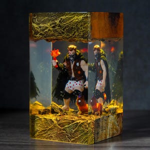 May include: A unique resin art piece featuring a muscular figure holding a flaming object, with a matching figure reflected in the resin. The figures are wearing a vest, cape, and heart-patterned underwear. A small cat figure is also present. The base has a textured, golden appearance.