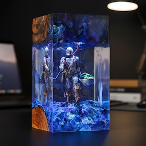 May include: A rectangular resin lamp featuring a Mandalorian figure carrying a small green creature in a pod. The base and top are made of natural wood, while the centre is clear resin with swirling blue and purple pigments and internal lighting.