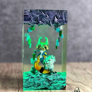 May include: A resin art piece featuring two fantasy characters in a clear rectangular block. The figures are in shades of blue, green, and yellow, set against a textured, glowing green base. The top of the block has a dark, rocky texture.