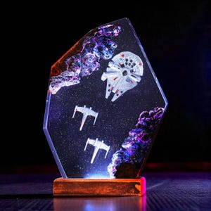 May include: A Star Wars-themed resin art piece featuring the Millennium Falcon and X-wing starfighters in a space scene. The artwork is set within a clear, geometric shape, mounted on a wooden base. The piece has a galaxy-like background with purple and blue hues.
