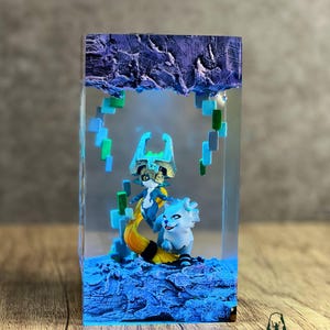May include: A resin sculpture featuring two fantasy characters in a blue and yellow color scheme, set within a clear rectangular block. The top of the block has a textured, purple appearance. The base is a textured blue. The sculpture is on a wooden surface.