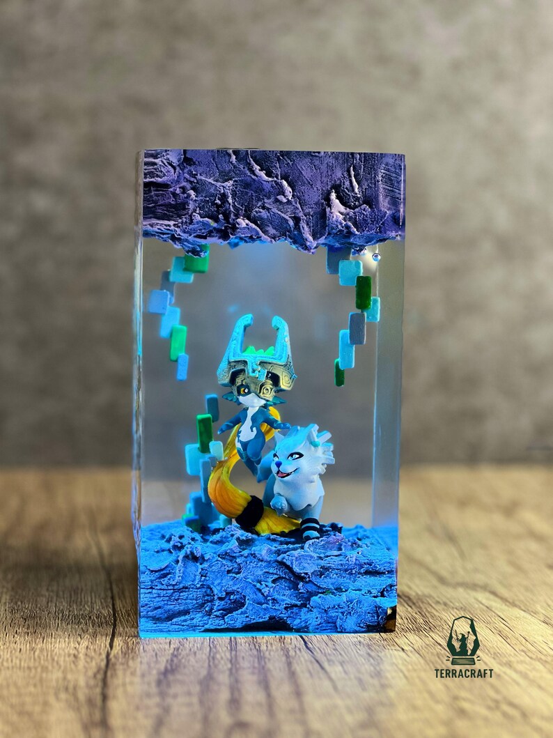 May include: A resin sculpture featuring two fantasy characters in a blue and yellow color scheme, set within a clear rectangular block. The top of the block has a textured, purple appearance. The base is a textured blue. The sculpture is on a wooden surface.