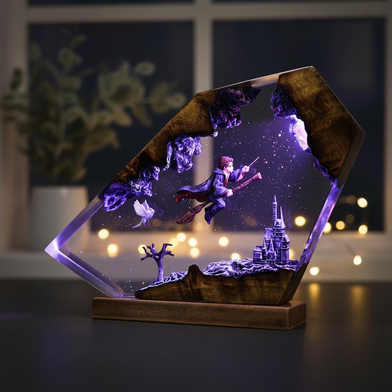 May include: A decorative lamp featuring a wizard on a broomstick flying over a castle, set within a clear, angled resin frame. The lamp has a wooden base and frame accents, with a purple and white color scheme. The scene is illuminated with a soft, purple glow.