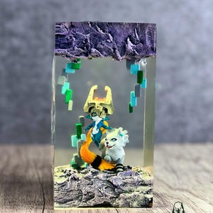 May include: A resin art piece featuring two figurines inside a clear block. One figurine is a blue and white character with a gold crown and orange tail. The other is a white creature with a black and white striped tail. The base is a textured, rocky surface.