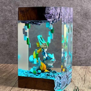 May include: A resin art piece featuring a blue and white creature with a figure on its back, set within a clear block. The base and top are made of wood and a textured purple material. The piece has a blue and green color scheme.