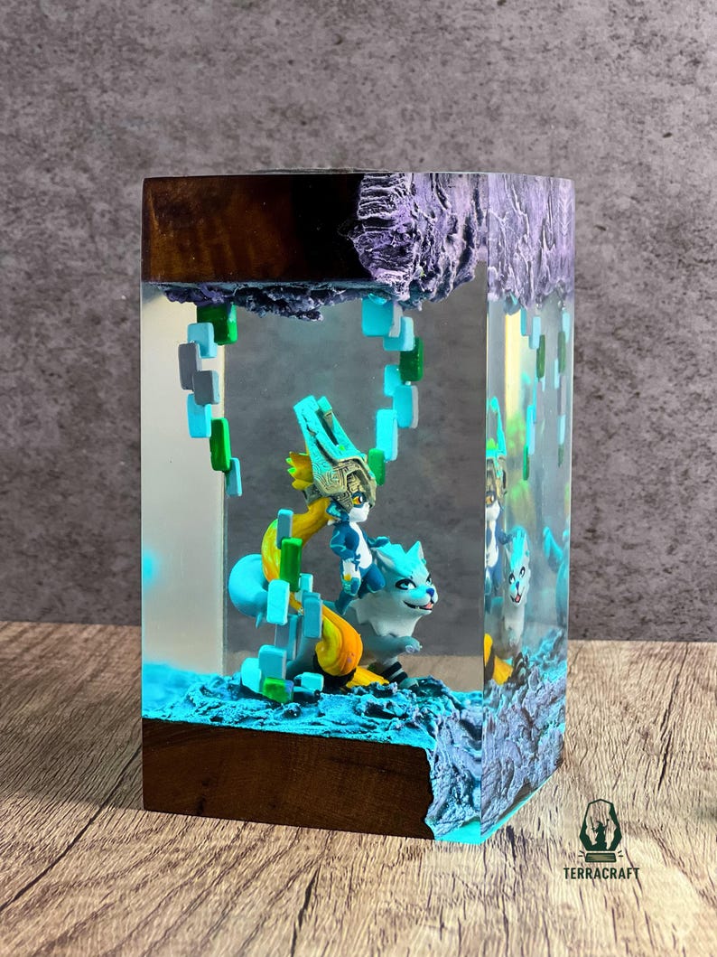 May include: A resin art piece featuring a blue and white creature with a figure on its back, set within a clear block. The base and top are made of wood and a textured purple material. The piece has a blue and green color scheme.