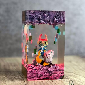 May include: A rectangular resin art piece featuring two fantasy characters inside. The base and top are textured, with a purple and pink color scheme. The characters are a blue and gold figure with a yellow tail and a white and pink creature.