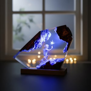 May include: A decorative resin lamp featuring a character holding a glowing star. The lamp has a wooden base and a clear, angled resin body with blue and brown accents. The scene is illuminated with blue light.