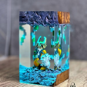 May include: A resin art piece featuring a fantasy scene with two white and yellow creatures, set within a clear block. The scene includes blue and green pixelated elements, and is set on a wooden base. The top of the block has a dark blue and gold textured design. The Terracraft logo is visible.
