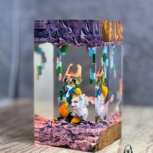 May include: A resin art piece featuring two figures riding white wolves, set within a clear block. The scene includes pixelated elements and a textured, purple base. The top has a gold and purple finish, with the "TERRACRAFT" logo visible.