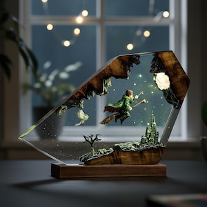 May include: A decorative resin lamp featuring a wizard on a broomstick, a castle, and an owl. The lamp has a wooden base and a clear, angled front. The scene is set against a starry night sky, with a glowing moon and lights.