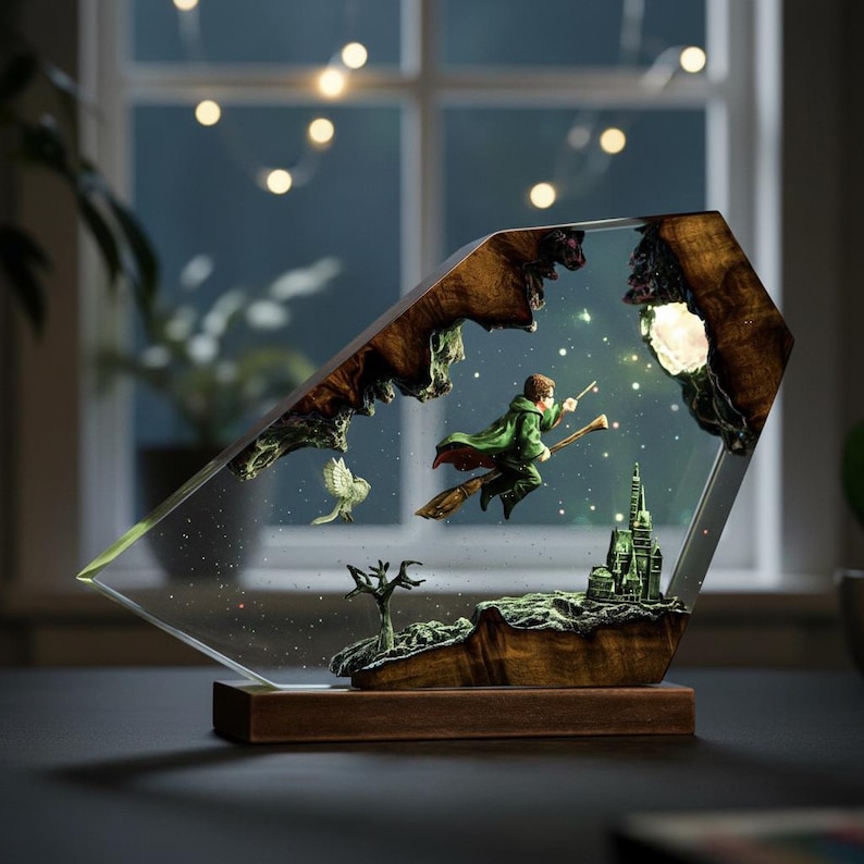 May include: A decorative resin lamp featuring a wizard on a broomstick, a castle, and an owl. The lamp has a wooden base and a clear, angled front. The scene is set against a starry night sky, with a glowing moon and lights.