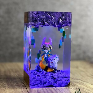 May include: A rectangular resin art piece with a wooden base, featuring two fantasy figures and a textured purple base. The figures are a fox-like creature with a long orange tail and a character with a purple mask. The piece is illuminated with blue light.