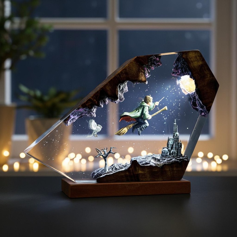 May include: A decorative resin sculpture featuring a wizard on a broomstick, an owl, and a castle. The sculpture is encased in a clear, angled frame with a wooden base. The scene is illuminated by a light source, creating a magical effect.