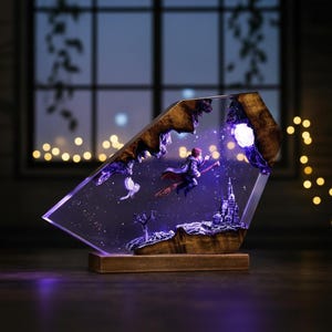 May include: A decorative resin sculpture featuring a figure on a broomstick, a castle, and a bird, set against a starry background. The sculpture is encased in a clear, angled frame with a wooden base and accents. The piece is illuminated with purple and white lights.