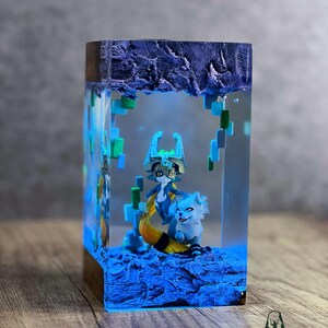 May include: A rectangular resin art piece featuring two fantasy creatures in a blue and green environment. The artwork is encased in clear resin with a wooden base and top. The piece is illuminated with blue light, creating a glowing effect.