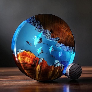 Könnte beinhalten: Ein runder Flaschenverschluss aus Holz mit einem blauen Harzdesign, das Mantarochen und einen Taucher in einem blauen Ozean zeigt. Der Verschluss hat ein dunkles Holzmaserungsmuster und einen kleinen, runden, dunklen Holzgriff.