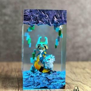 May include: A resin art piece featuring two fantasy characters inside a clear rectangular block. The characters are in shades of blue, white, and gold, set against a textured blue base and a purple, cloud-like top. The piece has a logo in the bottom right corner.