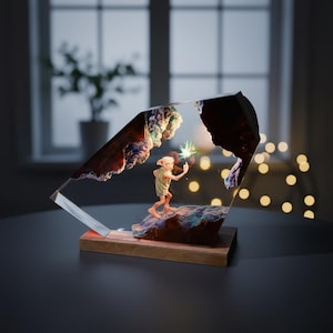 May include: A decorative resin sculpture featuring a character figure holding a glowing star. The sculpture is encased in a clear, angled block, set on a wooden base. The background shows a window and a string of lights.