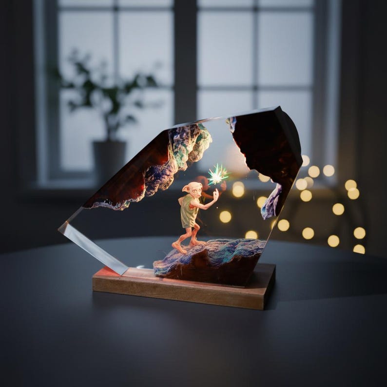 May include: A decorative resin sculpture featuring a character figure holding a glowing star. The sculpture is encased in a clear, angled block, set on a wooden base. The background shows a window and a string of lights.