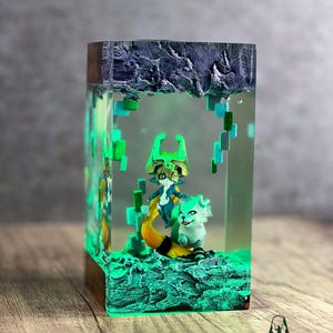 May include: A rectangular resin art piece featuring two fantasy figures inside a glowing green environment. The top and bottom are textured, with a wooden base. The figures are a fox-like creature and a character with a green mask and headdress.